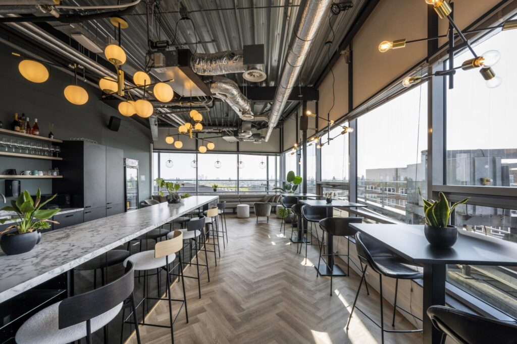 Modern lounge area at Bos en Lommerplein with high tables, bar seating, and large windows offering a city view.